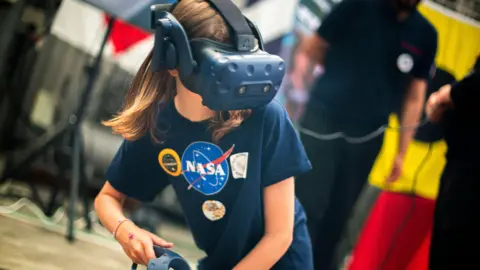 The picture shows a young person taking part in a virtual reality activity at what appears to be a science, technology or space‑themed event. In the centre of the image, the person is wearing a virtual reality headset that fully covers the eyes and upper face, with a thick strap running over the head. A handheld VR controller is visible in one hand. They are wearing a dark T‑shirt with a large NASA logo on the front, along with several circular patches or badges.