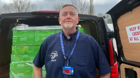 A man with a blue shirt looking into the camera and smiling. He has ginger hair and a ginger light beard. He is standing Infront of a van which is open that has piles of green boxes of easter chocolates. 