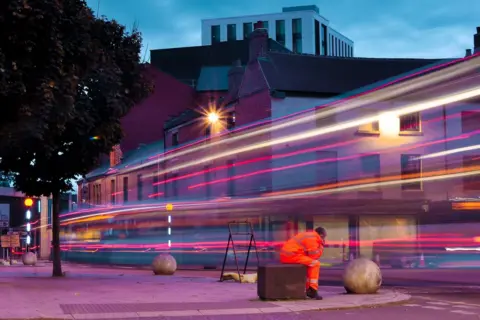 Neil J Catley An image of Coventry City Centre with a worker dressed in high-vis clothing sitting in the foreground and the lights of a vehicle seen drawn across the image