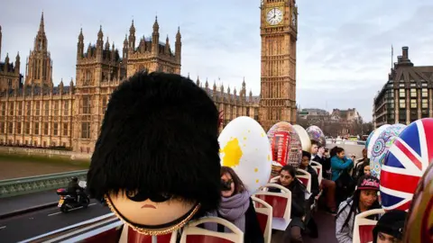 Nine large, decorated eggs, including one like like a beefeater and another the Union Jack, placed on seats on the roof of an open top bus with people sat in the seats, passing by the Houses of Parliament in the background. 