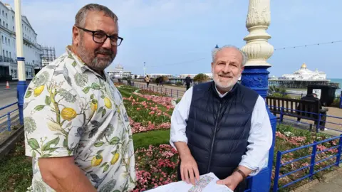 Lewes District Council and Eastbourne Borough Council Councillor Jim Murray poses for a photograph with landscape and garden designer Peter Thurman. Councillor Murray is wear a white, short-sleeved shirt featuring a design of yellow lemons on branches. Mr Thurman is wearing a navy gilet over a light blue shirt with rolled-up sleeves. They are standing in from of Eastbourne Carpet Gardens, blooming with pink plants. The sky is clear blue and Eastbourne Pier is visible in the background.