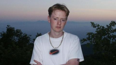 A man with red parted hair stands outside and poses for a picture during sunset. He is wearing a white top, a gold chain, and a gold watch. The top of a tree line can be seen in the background, as can hills faintly in the distance. The sky is a muted blue-grey colour with some dull orange.