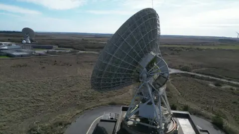 Goonhilly Earth Station The picture shows a very large parabolic satellite dish, also known as a radio or communications antenna, mounted on a heavy steel support structure. The dish surface is made from a metal lattice rather than a solid sheet, and it is angled upwards as if tracking signals in the sky. At the centre of the dish there is a feed horn and receiver assembly, held in place by metal struts that connect back to the main structure.