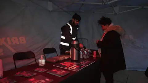Youth outreach team inside a large marquee tent, with a table and some leaflets laid out on it. There are also drinks, a hot drinks flask, and chairs behind one side. A woman stands on one side, and a man wearing a high vis jacket on the other