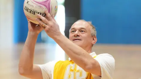 Ed Davey is about to try and shoot a netball ball. He is holding the ball up in front of his face. Mr Davey has short greying hair and wears a white polo t shirt with a yellow netball vest on with the letters GS on the front. He is holding a Gilbert netball ball which has pink swirls around the logo.