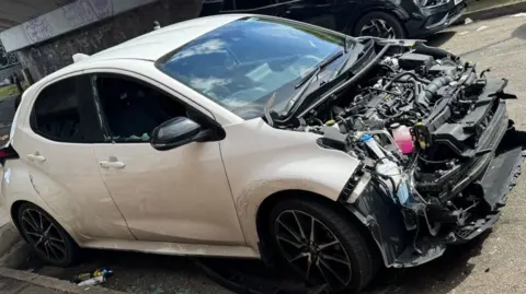 A white car with its bonnet lid missing and driver's side window smashed. It sits on a road under a concrete bridge which has graffiti on it.