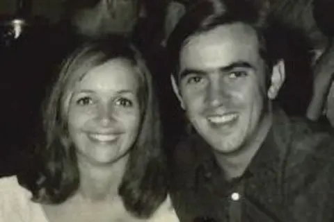 Rob Parsons Photograph of Rob (right) and Dianne (left) in their 20s they smile at the camera. Dianne smiles with her teeth at the camera and had brown shoulder length hair. Rob also smiles at the camera, has brown eyes and brown hair. It is a black and white image.