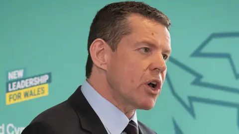 First Minister Rhun ap Iorwerth in black suit and gives a speech on stage