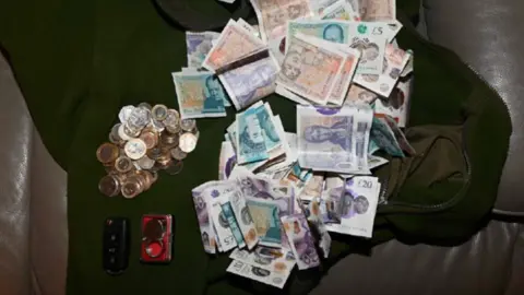 West Yorkshire Police Large amount of cash pictured on a dark sofa. Next to the cash are a pile of coins and a car key.