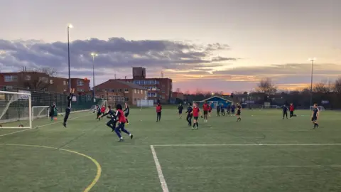 James Burridge/BBc Youngsters on a school astro pitch playing football. Buidlings visible in the background and floodlights on.