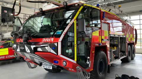 A large red fire engine is parked in a tender bay. The front has many windows - for all-round visibility. On its side there are two openings - where fire-fighting equipment can be seen. A smaller fire vehicle can be seen to the side - and a windowed door behind.