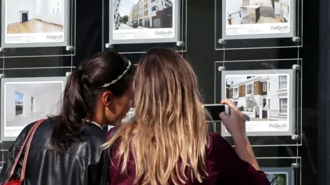 PA Media Two young women with their backs to the camera take a photo of an advert for a house in an estate agent's window. The woman on the left has black hair tied in a ponytail and a black leather jacket. The woman on the right has shoulder-length blonde hair and is holding her phone up to take a photo. 