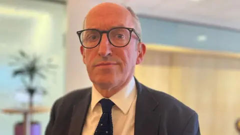 Mike McKirdy, who has receding grey hair and dark-rimmed glasses, looks at the camera in the close-up shot. He is wearing a dark suit, white shirt and blue tie. 