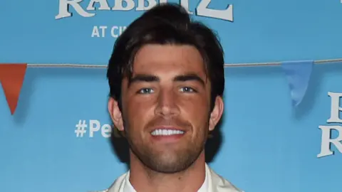 PA Media A man wearing a white shirt and jacket. He is at a movie premier and smiling as his picture is being taken.