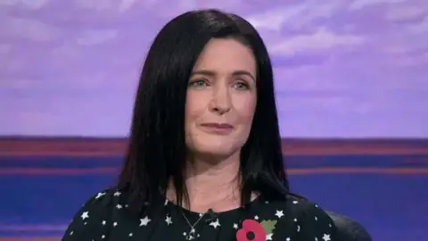 Paula Bradshaw with long dark, black hair is wearing makeup and a black top with white stars. Behind her the background is a mix of purple, green and orange.