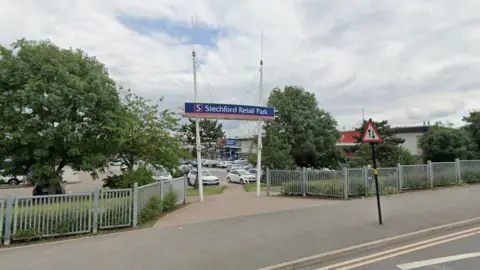 A Google street view image showing the entrance to a retail park on a pavement next to a main road. A blue, red and white sign above the entrance says "Stechford Retail Park". There are grey fences and trees either side. Beyond the entrance there is a car park and a line of large shops. 