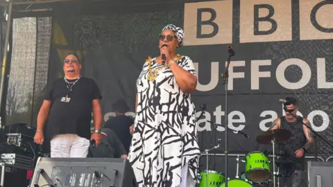 Ollie Ward/BBC A woman stands on a stage speaking into a microphone. She wears a long white and black top with white trousers, sunglasses and a white and black head cover.