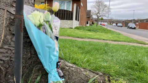 A bunch of white and yellow flowers leaning against a wall on a residential street. Houses on either side of the street and cars parked along the street.