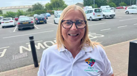 John Devine/BBC Nicola Christy has shoulder-length blonde hair parted in the middle. She has glasses and a white T-shirt, with the FACT logo on the right side. Behind her is a retail park, with lots of cars parked.