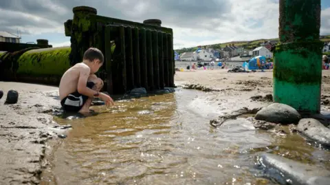 A boy in his shorts crouches in a stream of water on a beach next to a large mossy pipe with a grill on the end. In the distance a beach tent can be seen with people on the sand. 