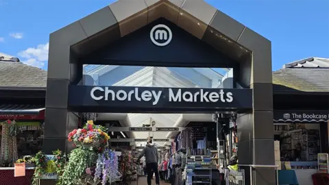 Chorley Council The entrance to Chorley Market. It shows a man from behind wearing a grey hoodie just inside the historic market looking at items on the stalls selling everything from books to clothes. The sky is blue with a few clouds.