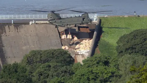 PA Media A Chinook helicopter with bags beneath hovering over a damaged reservoir wall