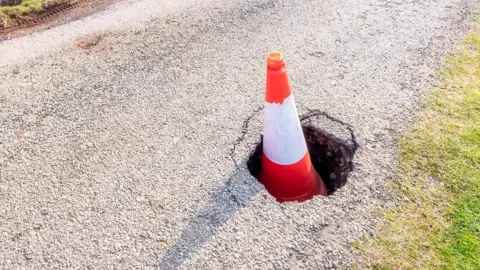 Getty Images A traffic cone sitting in a large pothole on a rural single-lane road.