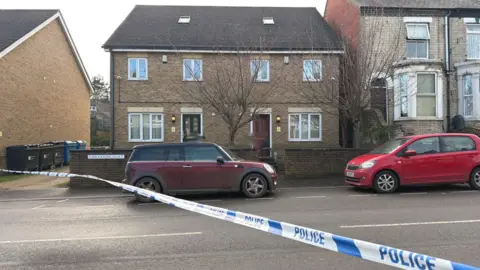 Richard Knights/BBC A modern-style semi-detached house has a red door open. Cars are parked outside and police tape stretches across the road.