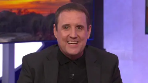 Peter Kay sits on a green sofa, he wears a black knitted polo shirt under a black jacket. He has short dark hair and smiles widely at the camera