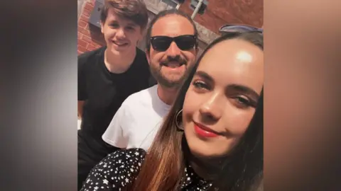 Handout A selfie of Mia with two friends. One has a beard and is wearing dark sunglasses and a white shirt, the other is wearing a dark t-shirt.