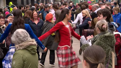 Dozens of people and dancers in Highland dress dance Strip The Willow on a street in Glasgow