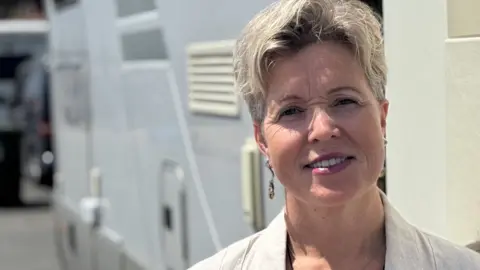 A head and shoulders shot of Katrina Cable. She is smiling and looking directly into the camera. She is wearing a cream shirt with a lace top beneath it. She is standing in front of her motorhome.