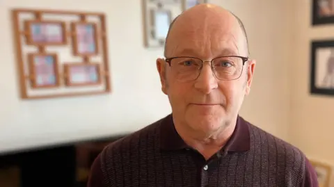 A bald man wearing glasses. He is standing in his living room looking directly into the camera.