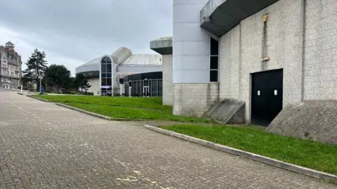 The outside of Plymouth Arena. There are two black doors on the right. The building is grey as well as the pathway leading to the entrance.