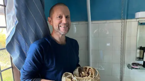 Fergus Drennan stands next to the toilet cistern at home holding a bag of acorns.