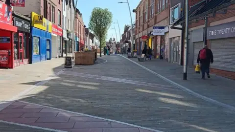 A high street which is lined with shops on either side. The shop in the foreground on the right is boarded up and there is a "to let" sign above the shop next to it. The pedestrianised street has planters and trees down the middle. There is a man walking away from the camera on the right and people visible in the distance.