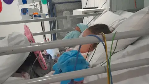 Samantha Hoodlass Myah lying in a hospital bed watching a tablet. She has various tubes and wires attached to her 
