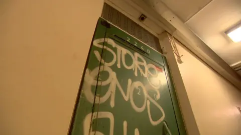 A shut green door to a flat in a corridor. The door has white writing sprayed upon it