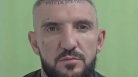 IOM CONSTABULARY Police mugshot of Thomas Lindop, who has close-cropped grey hair and a dark beard, and has Roman numerals tattooed on his forehead.