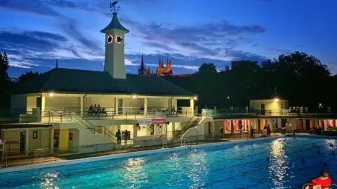 Vivacity Swimmers are in an open air pool that is bathed in moonlight and lighting around the pool. There is a two-storey building by the pool and people are watching from the terraces.