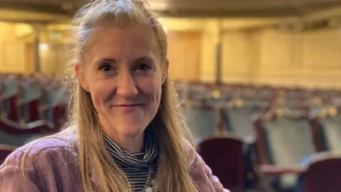 BBC Head-and-shoulders shot of Kerry Vasiliou, sitting in a theatre flooded with light. She is smiling and has long blonde hair tied back off her face.