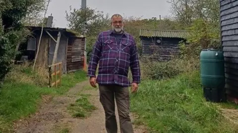 Richard Sanders, who has short grey hair and a beard, standing at the Othona camp. He is wearing a purple, checked shirt with brown trousers. He is surrounded by slightly overgrown grass and weary looking sheds. It is a grey day.