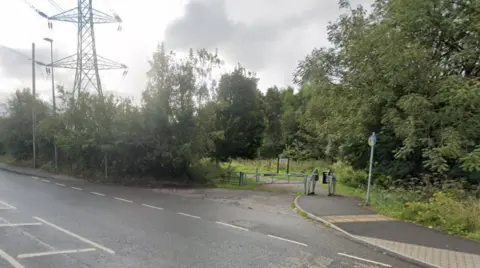 Google An entrance to the Linnyshaw Loopline and Ashtons Field in Little Hulton, Salford. A barrier to prevent vehicles blocks the path, which is surrounded on both sides by trees. 