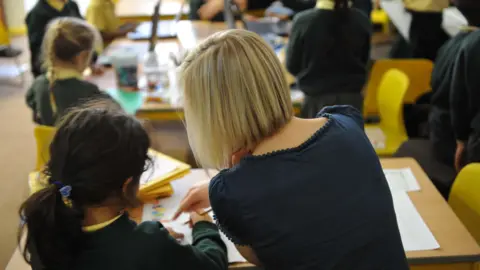 An anonymous girl in a school classroom being helped by an anonymous teacher. The picture shows them both from behind.