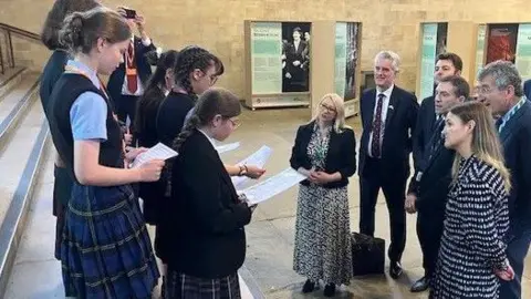 Andrew Sinclair/BBC Six school students stand on steps inside Parliament. Six Mps are gathered in front listening.