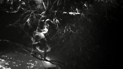 Durham Police A black and white image of a water rescue officer pulling one of the men to safety from the branch. The rescue officer has abseiled down on a rope. A lifebuoy floats in the water below them.