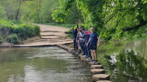 Epsom Trailblazers A group of man stand in a line on up-turned logs that run like stepping stones across a brown river.