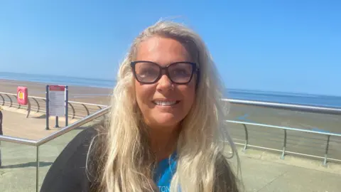 A closeup photo of a woman with blonde hair and black rimmed glasses, smiling to the camera in front of Blackpool Prominade