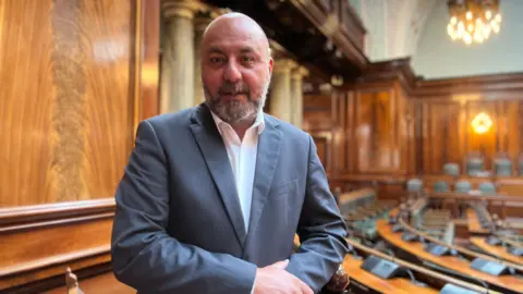 Aisha Iqbal/BBC A man in a grey striped suit and white shirt stands in an otherwise empty council chamber