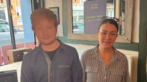 George King/BBC An Ed Sheeran cardboard cut out positioned next to Joe Gunter, a woman who is wearing a striped shirt and standing in a pub.